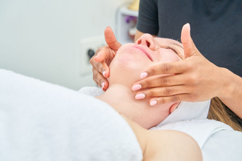 A woman is receiving a facial treatment from a woman. The esthetician slides her hands with oil over her neck.